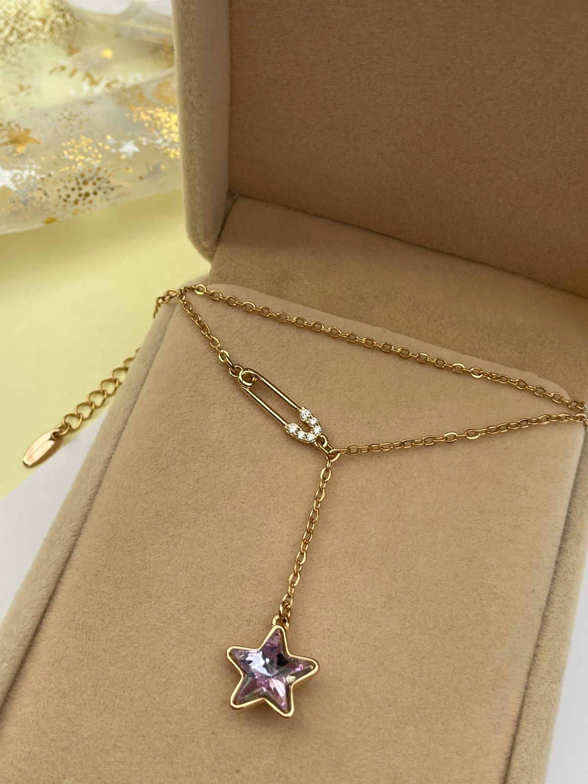 Collar dorado con estrella de cristal púrpura y pin de seguridad, mostrado dentro de su caja de presentación.