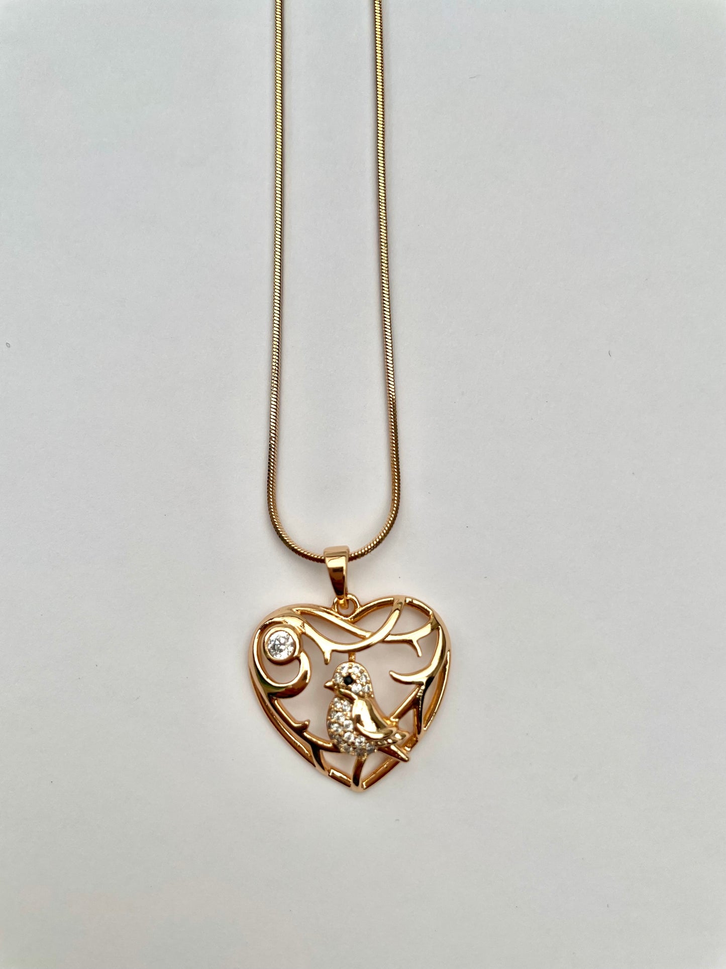 Collar dorado con cadena tipo serpiente y un dije en forma de corazón calado con un pájaro con incrustaciones de circones en su interior.