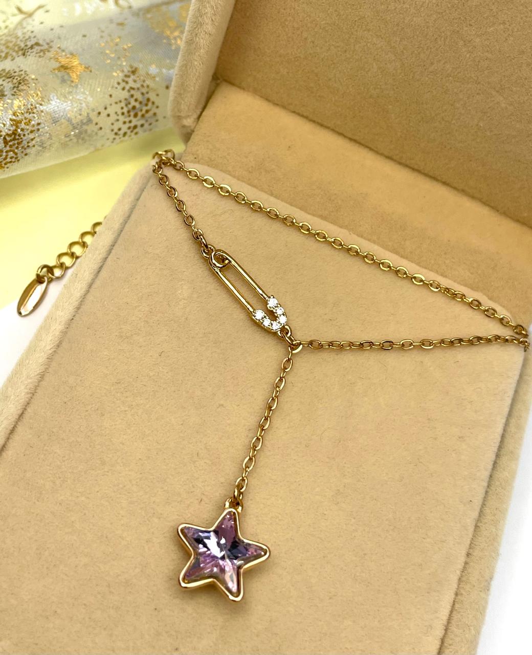 Collar dorado con estrella de cristal púrpura y pin de seguridad, mostrado dentro de su caja de presentación.