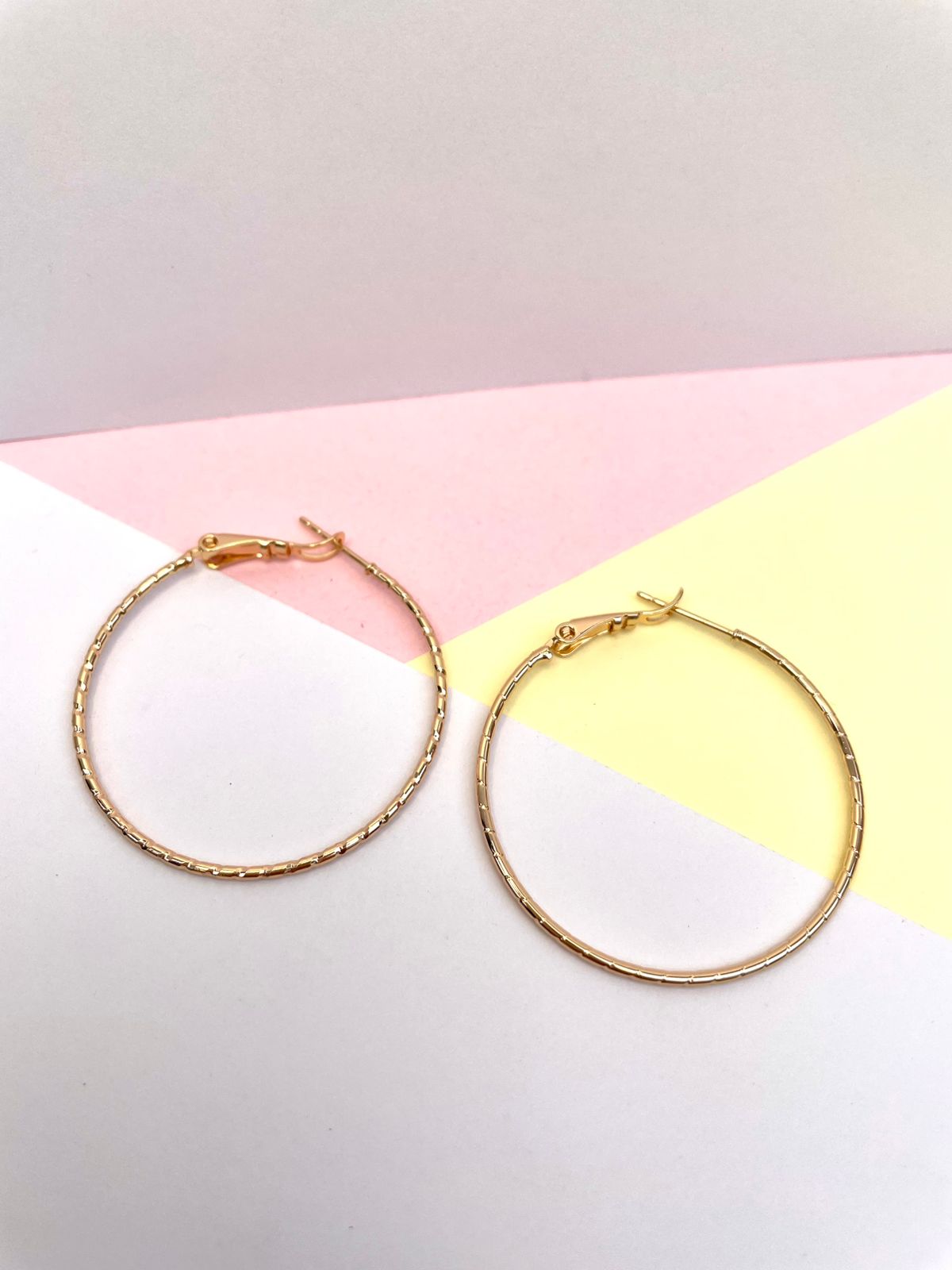 Par de argollas o aretes de aro delgados y dorados, con una superficie lisa y una textura tallada, sobre un fondo de colores pastel.