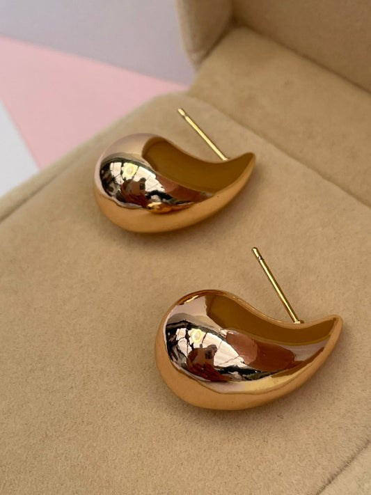 Par de aretes dorados con diseño escultural en forma de gota o coma, con un acabado mixto mate y pulido, dentro de una caja de joyería.