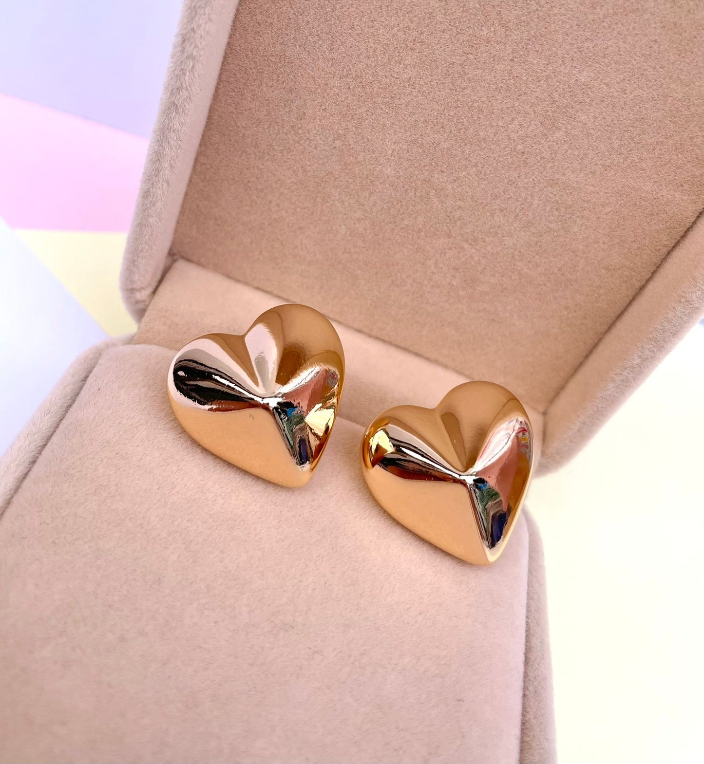 Par de aretes dorados en forma de corazón con volumen tridimensional y superficie lisa y pulida, dentro de una caja de joyería color beige.