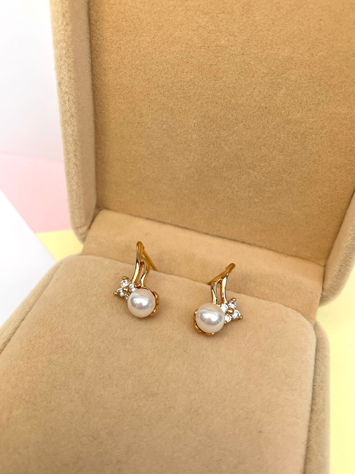 Par de aretes dorados con una perla de río central y tres pequeños cristales, presentados dentro de una caja de joyería color beige.