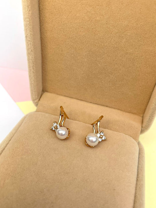 Par de aretes dorados con una perla de río central y tres pequeños cristales, presentados dentro de una caja de joyería color beige.