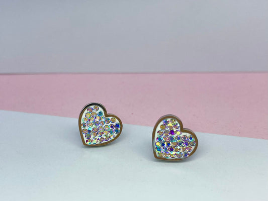 Par de aretes de stud en forma de corazón, cubiertos de cristales iridiscentes o tornasol, presentados de pie dentro de una elegante caja de joyería color beige.