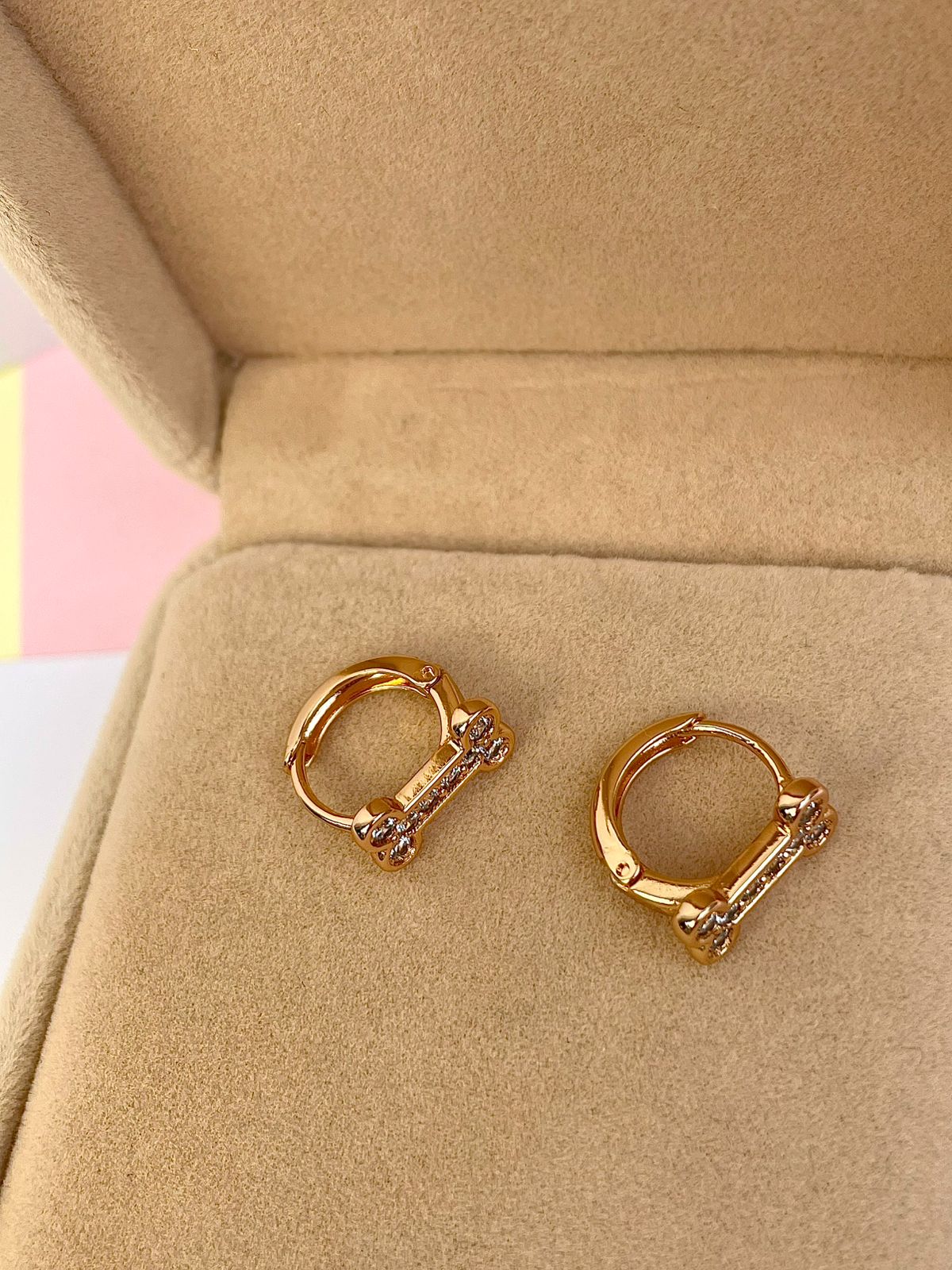 Aretes huggies dorados con diseño de huesito con cristales, presentados dentro de una elegante caja de joyería color beige.