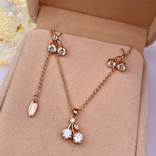 Set de joyas de collar y aretes con diseño de cerezas hechas de cristales brillantes, con un acabado en oro rosado y presentados en una caja de joyería.