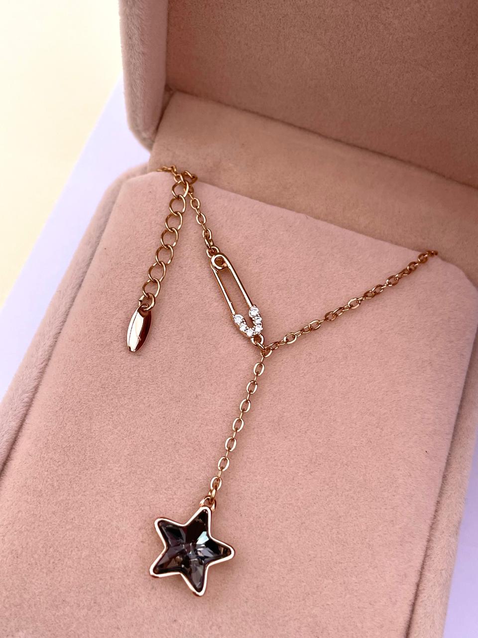 Collar dorado con estrella de cristal gris y pin de seguridad, mostrado dentro de su caja de presentación.