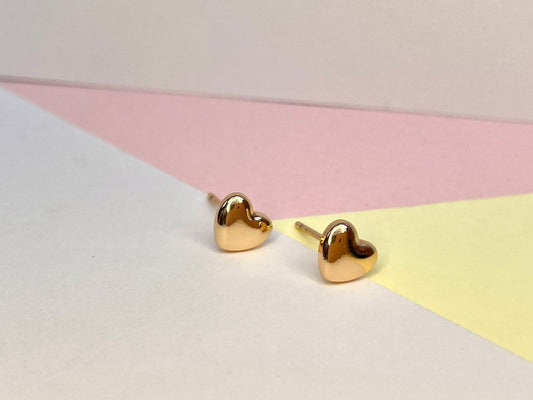 Par de aretes de stud dorados muy pequeños en forma de corazón, con un acabado pulido, sobre un fondo de colores pastel.