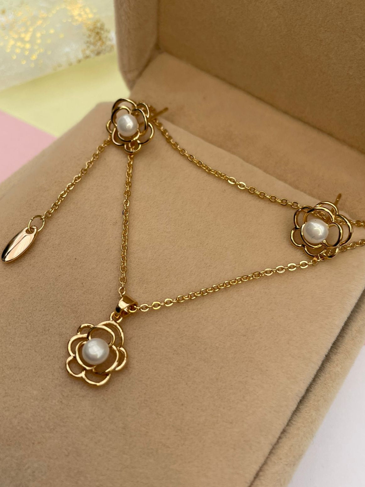Set de joyas de collar y aretes con diseño de flor dorada y perla central, presentado en una elegante caja de joyería color beige.