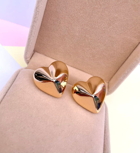 Par de aretes dorados en forma de corazón con volumen tridimensional y superficie lisa y pulida, dentro de una caja de joyería color beige.