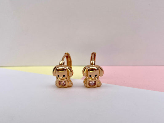 Par de aretes dorados en forma de perrito con piedra rosa en el pecho, sobre un fondo de colores.