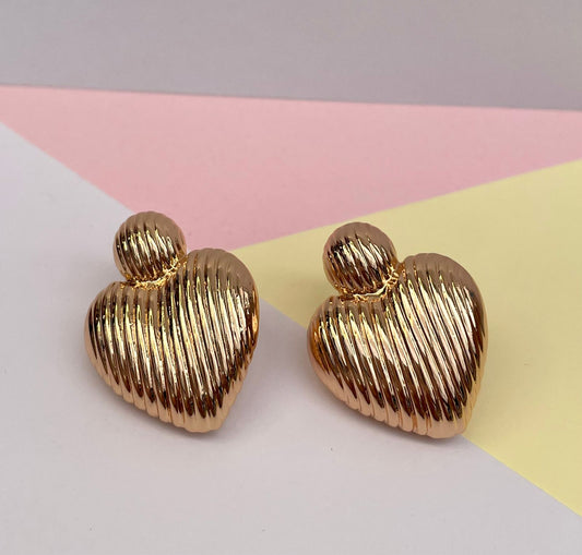 Par de aretes grandes dorados en forma de corazón con textura de líneas diagonales, presentados en una caja de joyería color beige.