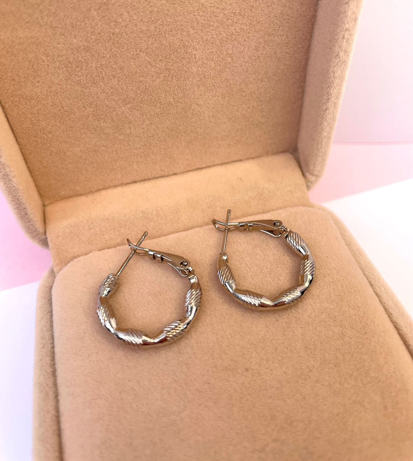 Par de aretes huggies pequeños de color plateado, con una textura tallada y entorchada que simula un corte diamante, dentro de una caja de joyería.