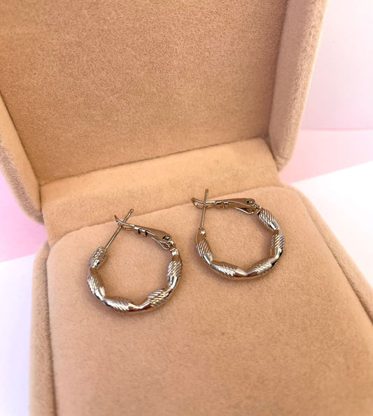 Par de aretes huggies pequeños de color plateado, con una textura tallada y entorchada que simula un corte diamante, dentro de una caja de joyería.
